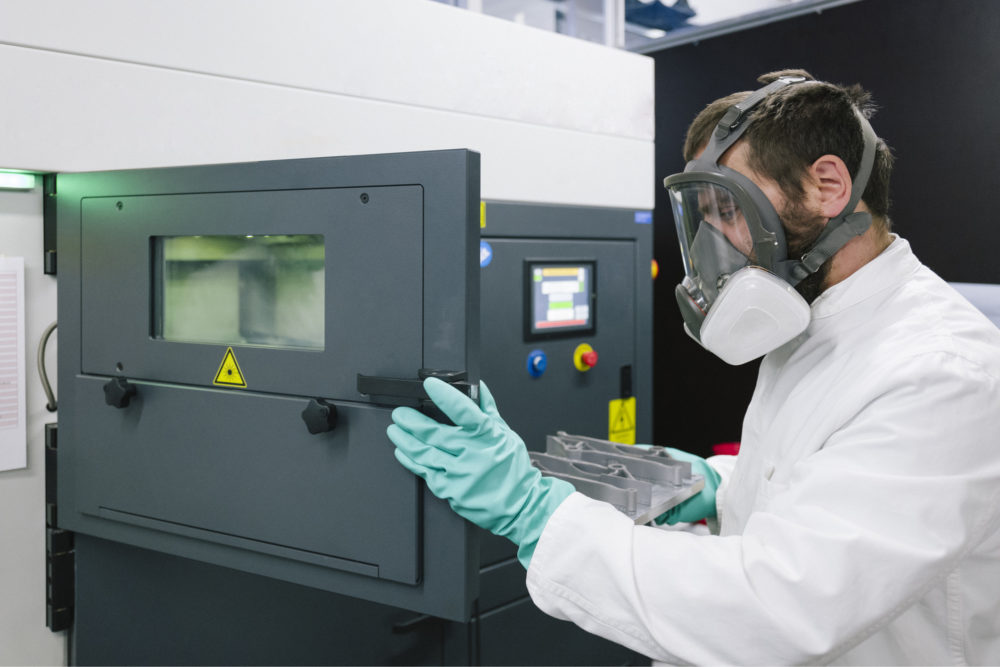 mężczyzna w masce przeciwpyłowej i fartuchu laboratoryjnym wyciągający wydruk z drukarki.