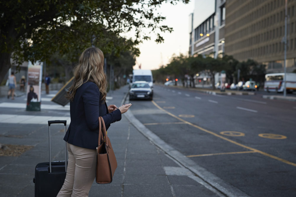 Businesswoman with shoulder bag hailing a cab with her phone, at dusk